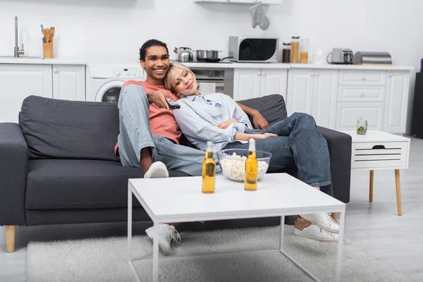 Jovem mulher loira se apoiando no homem americano africano feliz segurando controlador remoto na sala de estar — Fotografia de Stock