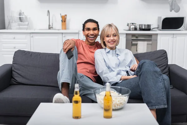 Feliz interracial pareja viendo película cerca de cerveza botella y palomitas de maíz en mesa de café - foto de stock