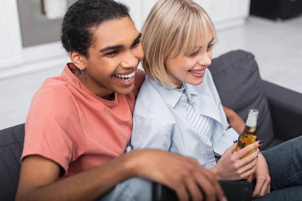 Vue grand angle de femme blonde heureuse tenant bouteille de bière près souriant homme afro-américain — Photo de stock