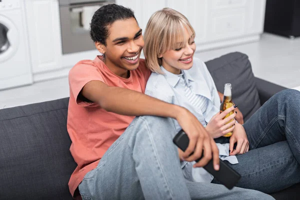 Felice donna bionda in possesso di una bottiglia di birra vicino sorridente uomo africano americano con telecomando — Foto stock