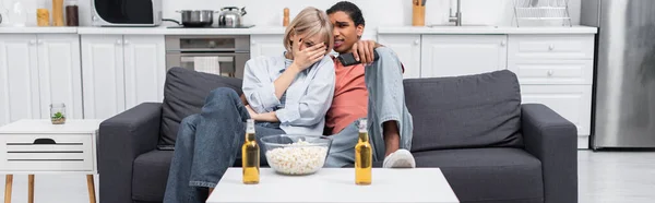 Young interracial couple watching scary movie in living room, banner — стоковое фото