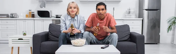 Joven pareja multiétnica animando mientras ve deporte partido en la sala de estar, pancarta - foto de stock