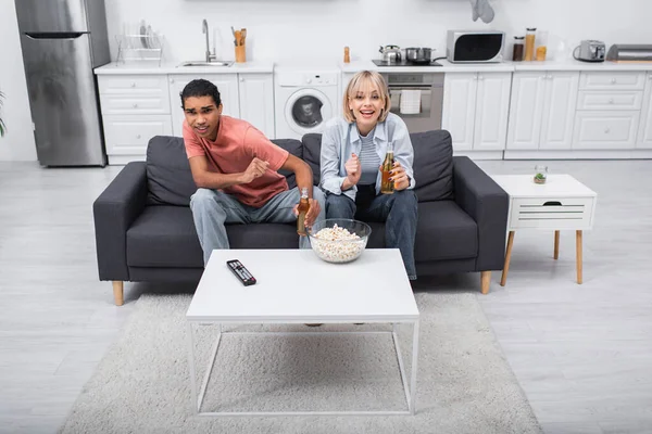 Emotional multiethnic couple cheering and holding bottles with beer while watching sport match in living room — стоковое фото