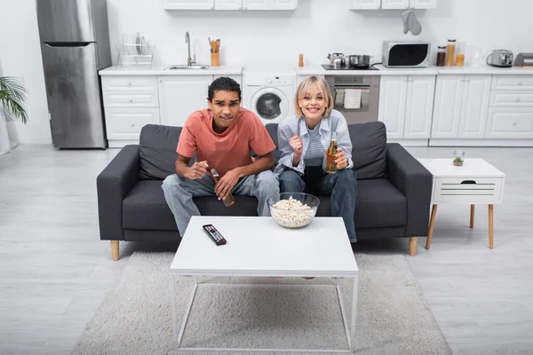 Emotional interracial couple cheering and holding bottles with beer while watching sport match in living room — Fotografia de Stock