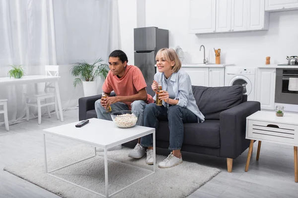 Young multiethnic couple holding bottles with beer while watching sport match in living room — Fotografia de Stock