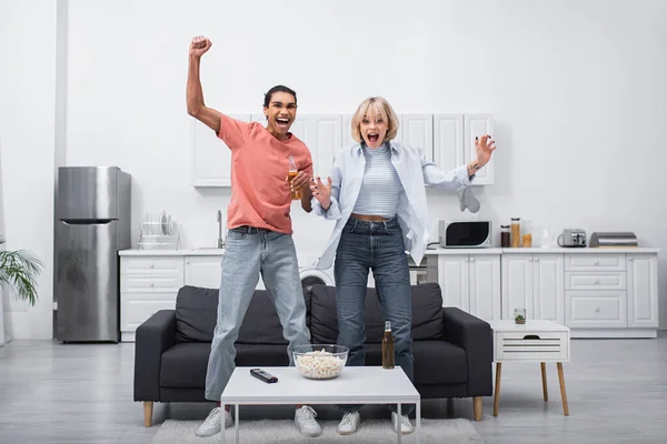 Animado casal multiétnico pulando enquanto assiste jogo esporte na sala de estar — Fotografia de Stock