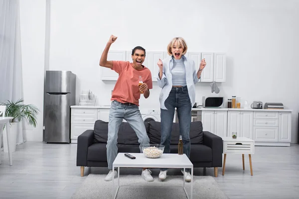 Feliz casal multiétnico pulando enquanto assiste jogo esporte na sala de estar — Fotografia de Stock