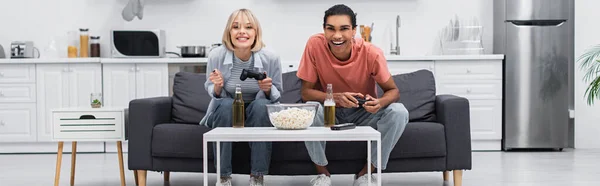 KYIV, UKRAINE - DECEMBER 6, 2021: happy multiethnic couple playing video game in living room, banner — стокове фото