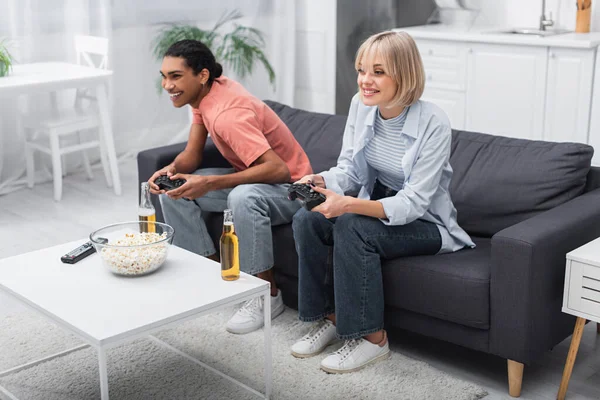 KYIV, UCRANIA - 6 de diciembre de 2021: feliz pareja multiétnica sosteniendo joysticks y jugando videojuegos - foto de stock