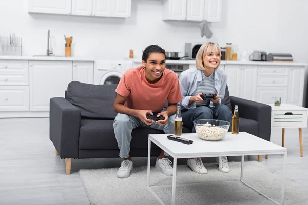 QUIIV, UCRÂNIA - DEZEMBRO 6, 2021: casal multiétnico alegre segurando joysticks e jogando videogame — Fotografia de Stock