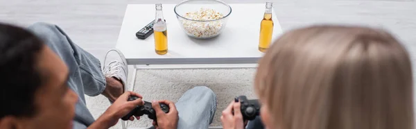 KYIV, UKRAINE - DECEMBER 6, 2021: popcorn and beer near multiethnic couple playing video game, banner — Stockfoto