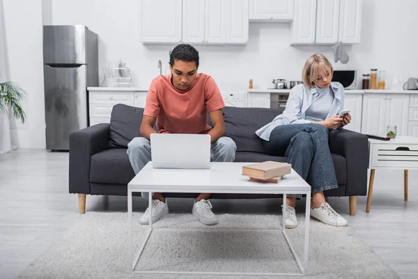 African american man using laptop near girlfriend with smartphone sitting on couch - foto de stock