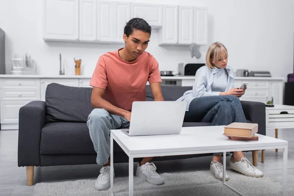 Homem americano africano usando laptop perto de namorada loira com telefone celular sentado no sofá — Fotografia de Stock