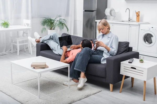 Homme afro-américain en utilisant un ordinateur portable avec écran vierge et couché près petite amie blonde avec téléphone portable sur le canapé — Photo de stock