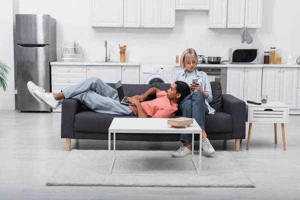 African american man using laptop with blank screen and lying near blonde girlfriend with smartphone on couch — стоковое фото