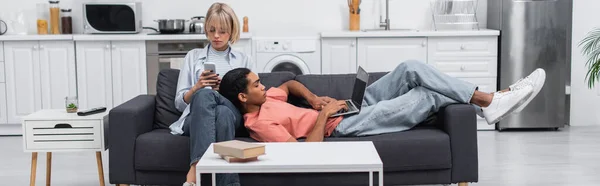 Hombre afroamericano usando portátil con pantalla en blanco y acostado cerca de novia rubia con teléfono inteligente en el sofá, pancarta - foto de stock