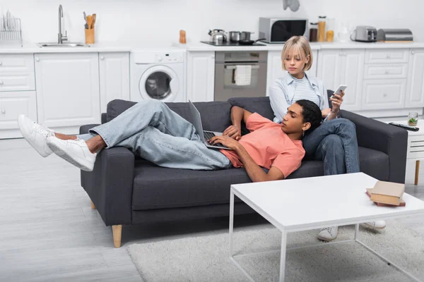 Blonde girlfriend holding smartphone and looking at african american man working on laptop — Stockfoto