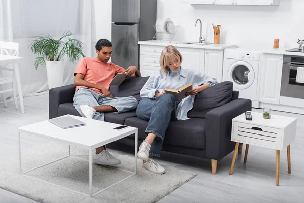 Young african american man and blonde woman reading books near gadgets in living room - foto de stock