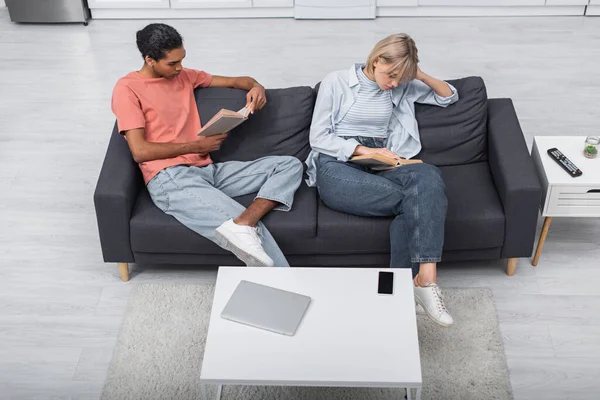 Vista superior de joven afroamericano hombre y mujer rubia leyendo libros cerca de gadgets en sala de estar - foto de stock