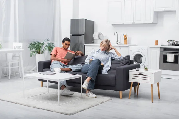 Jeune homme afro-américain et femme blonde lisant des livres près de gadgets dans le salon moderne — Photo de stock