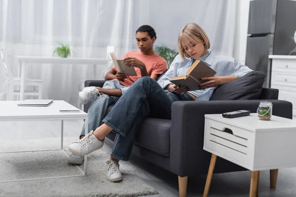Joven rubia y hombre afroamericano leyendo libros cerca de gadgets en la sala de estar moderna - foto de stock