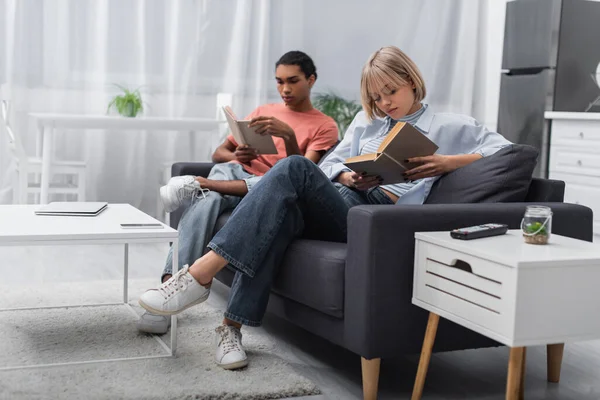 Young blonde woman and african american man reading books near gadgets in modern living room — Photo de stock