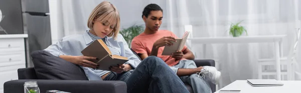 Young blonde woman and african american man reading books near gadgets in modern living room, banner — Photo de stock