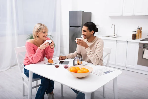 Счастливая многонациональная пара пьет чай во время завтрака и смотрит друг на друга — стоковое фото