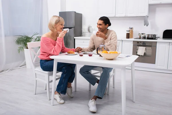 Heureux couple multiethnique boire du thé tenant la main pendant le petit déjeuner — Photo de stock