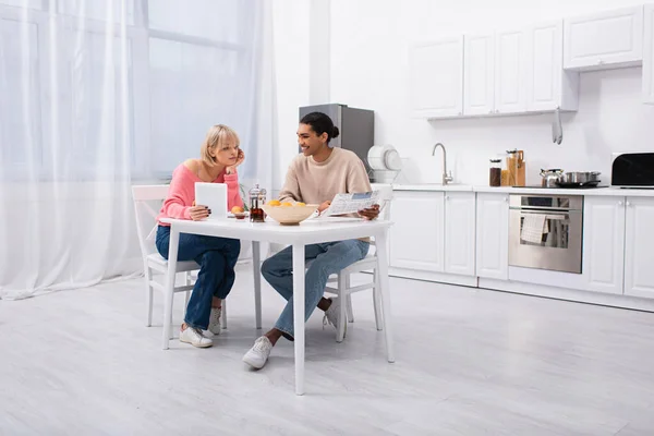 Heureux homme afro-américain tenant journal près de femme blonde avec tablette numérique pendant le petit déjeuner — Photo de stock