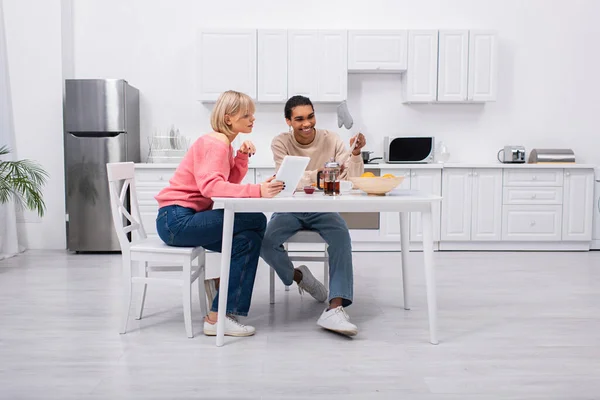 Cheerful african american man holding newspaper near blonde woman with digital tablet during breakfast - foto de stock