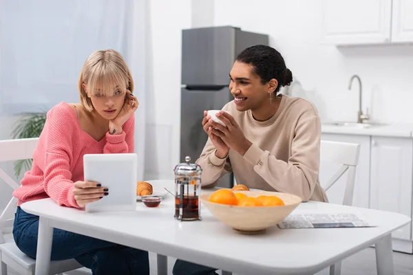 Happy african american man looking at blonde woman with digital tablet during breakfast — Foto stock