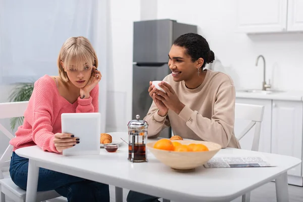 Happy african american man looking at blonde woman with digital tablet during breakfast — Fotografia de Stock