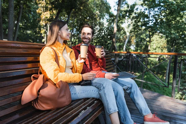 Turistas sorridentes com mapa segurando café para ir no banco ao ar livre — Fotografia de Stock