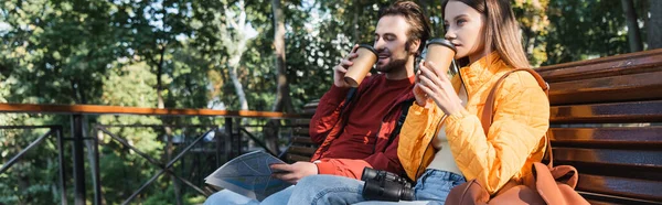 Viajero positivo sosteniendo mapa y café para acercarse a su novia en el banco al aire libre, pancarta - foto de stock