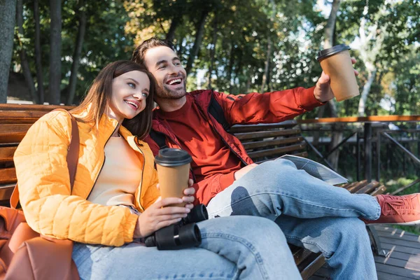 Усміхнений турист тримає каву, щоб піти і на карті біля дівчини з біноклями на лавці на відкритому повітрі — стокове фото