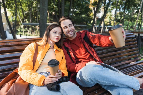 Viajero sonriente sosteniendo bebida para llevar y mapa cerca de la novia en el banco al aire libre - foto de stock