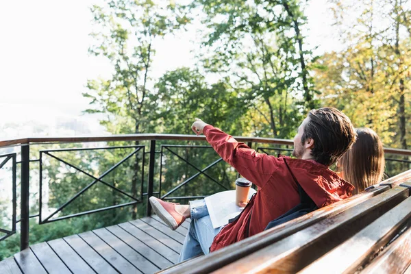 Young traveler pointing with finger and holding coffee to go near map and girlfriend on bench outdoors — Stock Photo