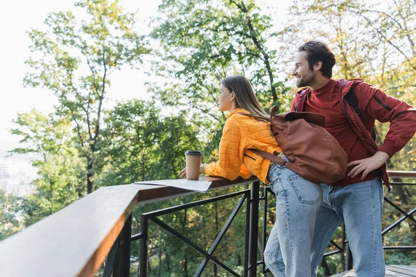 Vista lateral do viajante sorrindo olhando para longe perto da namorada com mochila, café para ir e mapa ao ar livre — Fotografia de Stock