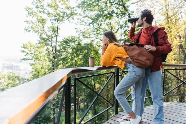 Side view of traveler looking through binoculars near girlfriend, map and coffee to go outdoors — Stock Photo