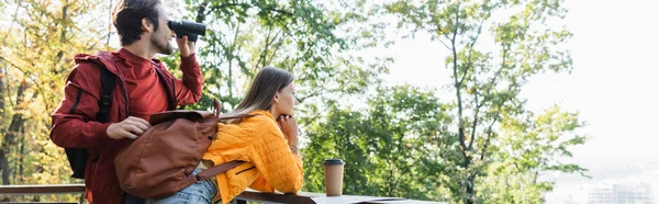 Vista lateral do turista olhando através de binóculos perto da namorada com mochila, café e mapa ao ar livre, banner — Fotografia de Stock