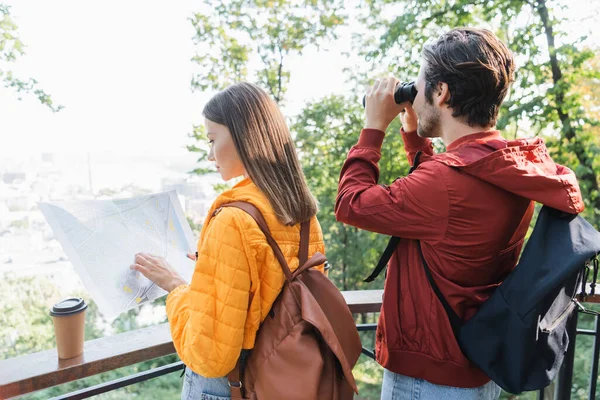 Vista lateral da jovem olhando para o mapa perto de café para ir e namorado com binóculos ao ar livre — Fotografia de Stock