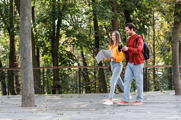 Cheerful couple of travelers holding binoculars, map and coffee to go on urban street — Stock Photo