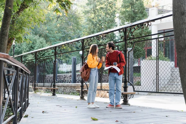 Side view of positive travelers holding coffee to go and map while talking on alley — Stock Photo