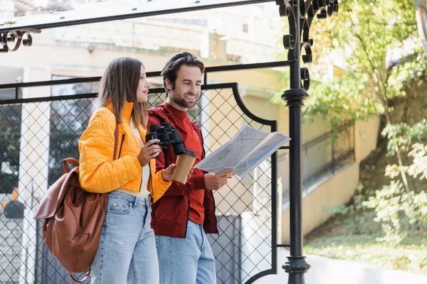 Positive tourist holding map near girlfriend with coffee to go and binoculars on urban street — Stock Photo