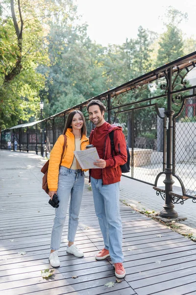 Positive tourists with binoculars, map and backpacks looking at camera on urban street — Stock Photo