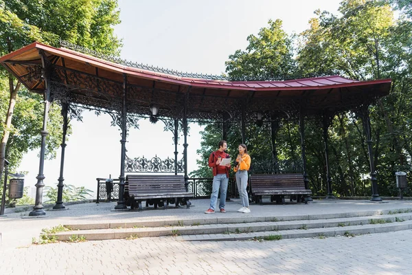 Smiling travelers with coffee to go and binoculars talking to boyfriend with map on urban street — Stock Photo