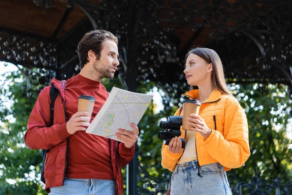 Turismo sosteniendo mapa y café para acercarse a su novia con prismáticos al aire libre - foto de stock