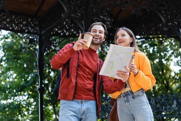 Low angle view of smiling tourist holding paper cup and map while pointing with finger near girlfriend outdoors — Stock Photo