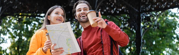 Low angle view of cheerful man holding map and pointing with finger near girlfriend with coffee to go on urban street, banner — Stock Photo